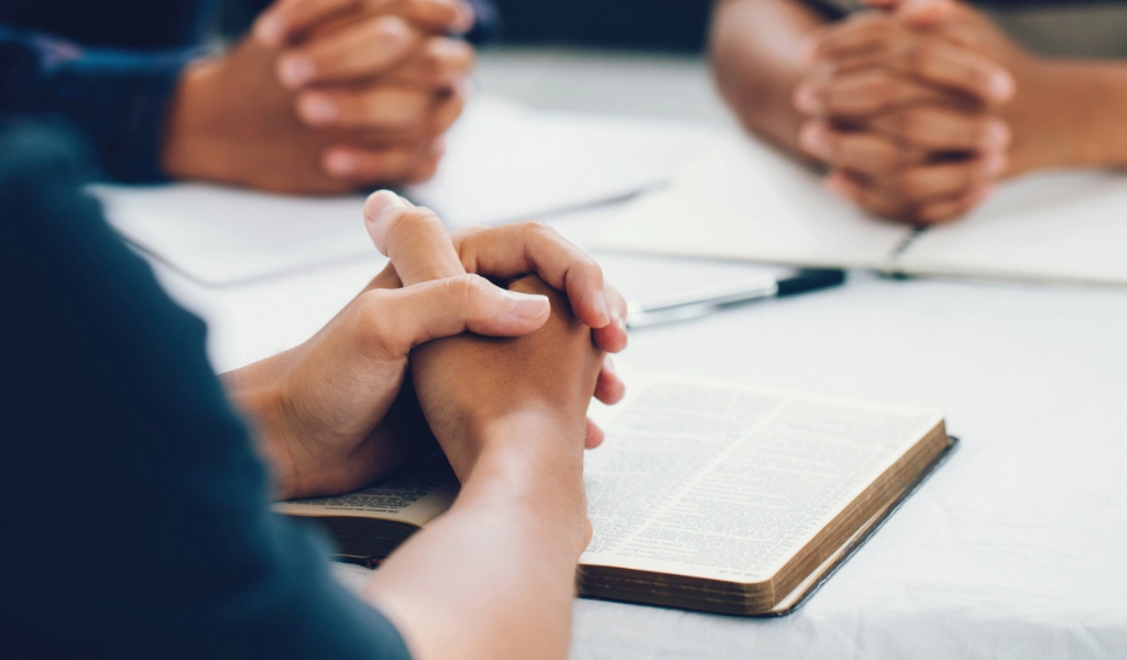 group of people studying with folded hands on a bible.